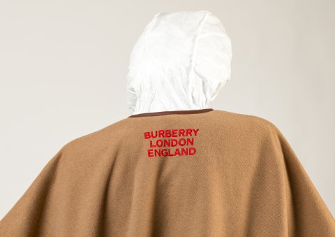 Rear view of mannequin's head and shoulders, wearing a white hood and brown cape embroidered with BURBERRY LONDON ENGLAND.